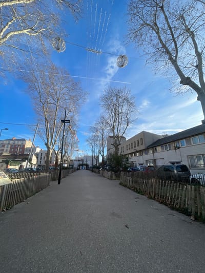 The image depicts a paved pedestrian walkway in Gennevilliers, France, under a clear, bright blue sky with scattered thin white clouds and a prominent aircraft contrail extending horizontally. The time of day is sunny, likely late morning or early afternoon.
The walkway, constructed of dark gray asphalt or pavement, extends from the foreground into the distance, narrowing in perspective. It is flanked on both sides by low, rustic wooden fences made of vertical stakes, separating the path from adjacent areas.
Overhead, a network of white festive string lights is visible, stretching across the pathway between bare deciduous trees. Two large, spherical, open-frame decorative globes, possibly illuminated, hang from these overhead wires. Numerous leafless trees, characteristic of late autumn or winter, line both sides of the path, with their branches reaching into the sky.
On the left side of the path, behind the wooden fence, an elevated section of ground with a retaining wall is visible, followed by a street or parking area with several parked cars. Further back, multi-story urban buildings, including one with a reddish-brown facade, rise above the street level. A dark street light pole stands prominently on the left side of the path. Partially visible in the extreme foreground on the left is a brown interpretive sign or display board.
On the right side of the path, beyond the wooden fence, a parking area contains multiple vehicles, including a dark-colored van or SUV. Adjacent to this parking area is a multi-story building with a light-colored facade and numerous rectangular windows, suggesting an administrative or residential structure. Another taller building is visible further down the path on the right.
The path recedes towards an opening in the mid-distance, flanked by buildings, suggesting a continuation into another section of the urban environment or a public square. No individuals are present within the frame.
