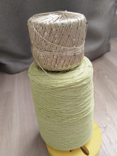Two spools of thread are stacked on top of each other. The bottom spool is larger and a light green color. The top spool is smaller, a tan color, and decorated with small, silver sequins. The spools are sitting on a light-colored wooden surface. In the background, a gray fabric is visible.