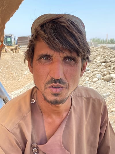 A head-and-shoulders, close-up portrait of a man is depicted outdoors. The man has tan skin, dark, tousled hair, and a short, dark beard. He is wearing a light brown, loose-fitting garment with a band collar and metal snap closures. On his head, he wears a traditional, woven cap. His eyes are dark and direct, looking at the camera. The background consists of dry, rocky terrain with a construction vehicle partially visible on the left, and piles of rocks and debris on the right, under a bright, clear sky. The overall impression is one of a candid, possibly work-related, outdoor setting in a dry, arid environment.