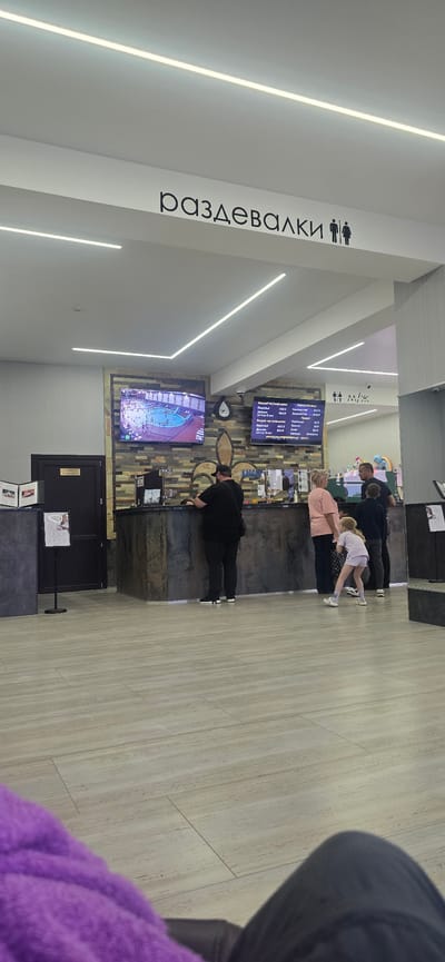 The image depicts an indoor scene, likely a changing room area or reception in a facility in Suvorovskaya, Russia. The dominant text on a white pillar reads "РАЗДЕВАЛКИ" which translates to "CHANGING ROOMS", with male and female restroom icons above it.

Several people are visible. In the foreground, a person wearing a purple fuzzy garment is partially visible, facing away from the camera. Further back, a man in a black shirt and pants, wearing a baseball cap, is leaning over a counter or desk. To his right, a woman in a pink shirt and dark pants is standing, facing the counter. Next to her, a young blonde person in a white top and patterned shorts is bent over, as if doing a physical activity or being coached. Behind them, a man in a dark checkered shirt is visible.

The setting appears to be modern, with a light-colored wooden floor and sleek white walls. Linear LED lights are integrated into the ceiling, providing illumination. There are at least two large screens displayed on the wall behind the counter. One screen shows what appears to be a live video feed of an indoor area with multiple people, possibly a sports facility or entertainment venue. The other screen displays a menu or price list, with text in Russian. A dark door with a sign indicating "КАМЕРА ХРАНЕНИЯ" (STORAGE ROOM) is visible. The overall atmosphere seems casual and functional. The time of day is not discernible from the image.