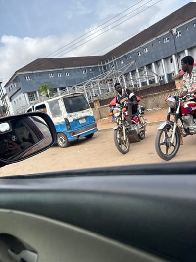 Two young men are riding motorcycles on a street in Obinze, Nigeria. The man in the foreground is wearing a floral shirt and a black patterned vest over a white shirt. The man behind him is wearing a black and white vest with "Michigan" written on it, over a white shirt. Both men are wearing dark pants. They are riding on a dusty road in front of a blue and white van. In the background, there is a large, modern-looking building with a dark roof and many windows. The sky is overcast with some clouds. A side mirror of a vehicle, likely a car, is visible in the lower left corner of the image, showing a reflection of the interior of the car and the sky.