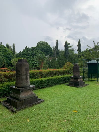 Two stone pillars, likely of a religious or historical significance, are situated in a park-like setting in Mungkid, Indonesia (latitude: -7.604, longitude: 110.23). The pillars are erected on tiered stone bases, and the area surrounding them is covered in lush green grass and manicured hedges. The pillars appear to be made of rough, grey stone, suggesting age and natural weathering. In the background, a small green building, possibly a restroom or a kiosk, is visible. The scene is framed by a cloudy sky, suggesting a recent rain shower. The overall setting exudes a sense of tranquility and historical significance.