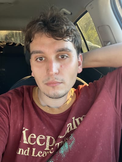 A medium shot shows a young man with dark, messy hair sitting in the driver's seat of a car, looking at the camera. He is wearing a maroon t-shirt with a yellow collar and a faded gold design that reads "Keep Calm and Leave." His left arm is resting behind his head. The background shows the interior of the car and a blurred view of greenery and a car through the window, suggesting an outdoor location. The image is a selfie taken from a slightly low angle.
