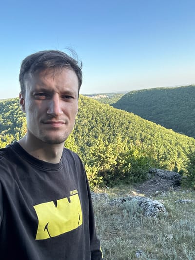 The image is a selfie taken by a man with short, dark hair and a light complexion. He is wearing a dark, long-sleeved t-shirt with a large yellow graphic on the left side and text that reads "THE ORIGINAL". He is positioned in the foreground, slightly to the left of center.
The background depicts a vast, lush green landscape of rolling hills covered in dense forest. The sun is setting or rising, casting a warm light on the scene and creating long shadows. The sky is clear and blue. In the distance, on the left side of the frame, a small town or village can be seen nestled among the trees. The terrain in the foreground where the man is standing is rocky and covered in dry grass and low-lying vegetation. The overall impression is one of a scenic, natural environment.
The image appears to have been taken in the vicinity of Pryvorottia Druhe, Ukraine, given the landscape's characteristics which are consistent with the region's geography.