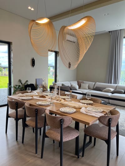 The image depicts a modern, well-lit dining area in a home. The focal point is a wooden dining table set for a meal, adorned with plates, glasses, cutlery, and a runner. Two distinctive, large, artistic pendant lights hang above the table, casting a warm glow.
The dining table is surrounded by upholstered chairs with black legs. In the background, a large, modular sofa with numerous cushions is visible, suggesting a comfortable living space adjacent to the dining area. Large windows offer views of a green lawn and a suburban landscape, indicating a location outside of a dense urban core. A hint of the location, Yakunino, Russia, is suggested by the presence of trees and houses in the distance consistent with a Russian dacha or suburban environment. The overall ambiance is one of contemporary elegance and domestic comfort.