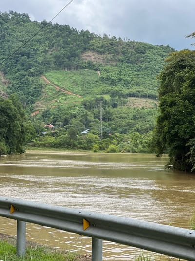 A wide, muddy river occupies the foreground and midground of the image, reflecting the overcast sky with a shimmering, textured surface. On the far bank of the river, a lush green, densely forested hillside rises steeply, with patches of cleared land and a winding dirt path visible on its slopes. Scattered amongst the trees on the hillside are several houses with red and grey roofs, suggesting a small village or settlement. A tall, grey metal electricity pylon stands prominently on the mid-slope, connected to overhead wires. The riverbanks are lined with thick, green foliage. In the lower foreground, a silver metal guardrail with yellow reflectors runs horizontally across the frame, partially obscuring the view of the river and its grassy bank. The scene suggests a rural or semi-rural setting in or near Hà Giang City, Ward 2, Vietnam, possibly after recent rainfall given the muddy water.