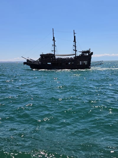 A dark-colored, two-masted pirate-style ship is sailing on the Aegean Sea in front of Thessaloniki, Greece. The ship has a wooden deck with several people visible, some standing and some seated. It is a sunny day with a clear blue sky and gentle waves on the sea, reflecting the sunlight. The ship's hull is dark, and it features characteristic pirate ship elements like cannons and a prominent bowsprit. In the distance, a hazy shoreline with faint mountains can be seen, suggesting the proximity to the city of Thessaloniki. Another smaller boat is visible in the far distance. The water is a vibrant turquoise color.