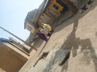 The image displays a daytime street scene in Gobirawa, Nigeria, captured from an elevated, oblique perspective looking down a thoroughfare. Three individuals, likely adult females, walk away from the camera along a dusty, uneven ground surface that appears to be cracked pavement or packed earth. The individual closest to the viewer wears a purple full-length garment; the middle individual wears a white full-length garment, and the furthest individual wears a yellow full-length garment. All three wear head coverings. Strong sunlight from the upper left casts prominent shadows of the individuals and a utility pole onto the ground. Buildings line both sides of the street. On the right, structures include a light-colored multi-story building with multiple windows, and further down, a building featuring a yellow and black painted section with the number "101" visible. On the left, a wooden utility pole stands with overhead wires, and simpler, low-rise buildings with unpainted brick walls are present. The ground shows signs of wear, cracks, and scattered debris.