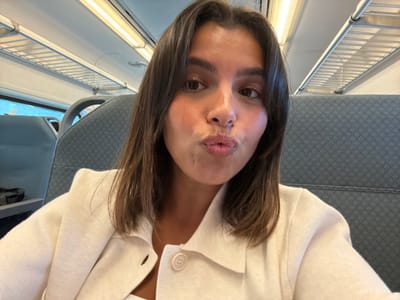 A young adult female is depicted in a close-up selfie within a train carriage. She has medium-length dark hair and brown eyes, looking directly at the camera with her lips pursed in a "kiss" pose. She is wearing a light-colored, possibly off-white, collared cardigan or jacket over a lighter top. The background reveals typical train interior elements: patterned grey fabric seats with headrests, a metal slatted overhead luggage rack, and fluorescent strip lighting on the ceiling. A window is partially visible on the left, showing diffused exterior light. The scene is located within a train carriage, in the city of Linden, United States.