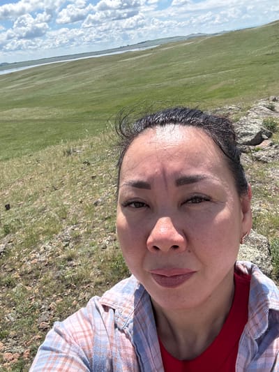 A woman is taking a selfie in a grassy, hilly landscape. She is wearing a red shirt and a blue and pink plaid shirt. Her hair is dark and pulled back. The background shows rolling green hills, a body of water, and a cloudy sky. The terrain in the foreground is rocky and covered with dry grass. The location is the city of Aumyshevo, Russia.