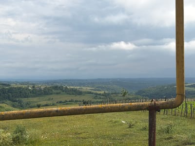 A horizontal, yellow, rusted pipeline stretches across the foreground of the image, supported by a vertical, rusted pipe in the center. The pipeline is positioned in front of a lush, green landscape of rolling hills dotted with trees and scattered buildings. A low, rustic fence made of wooden posts is visible behind the pipeline, separating the grassy field from the more distant terrain. The sky above is overcast with soft, gray clouds. The scene appears to be located in Kalauri, Georgia, characterized by its hilly topography and rural setting.