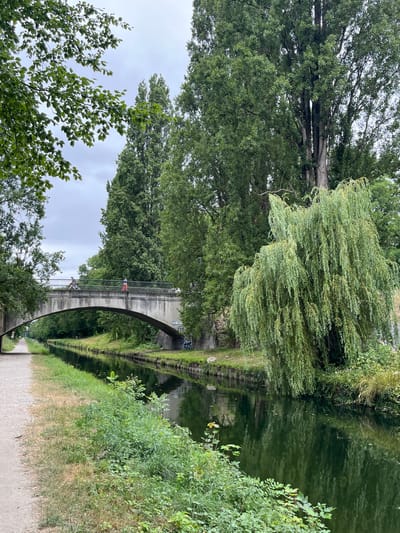 An overcast day in Villeparisis, France, features a canal with a concrete bridge spanning it. A dirt path runs along the left side of the canal, with lush green trees and foliage lining both banks. On the bridge, two individuals are visible; one is on a bicycle and the other is walking. One person is wearing a blue top and dark bottoms, while the other is wearing a red top and dark bottoms. The water of the canal reflects the surrounding trees and sky. A prominent weeping willow tree stands on the right bank, its branches cascading towards the water. The scene appears peaceful and serene, showcasing a natural urban landscape.