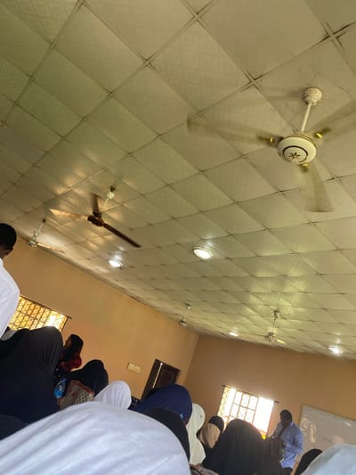 The image shows an interior scene, likely a mosque or place of worship, in Sabo, Nigeria. The ceiling is tiled in a diamond pattern, with several ceiling fans visible; one is white with gold accents, and another is dark brown. Recessed lighting is also present. Several people are present, most likely in a prayer position. They are wearing various clothing items, including white head coverings, black garments, and blue attire. Light enters the room through windows. A whiteboard is partially visible in the upper right corner of the image.