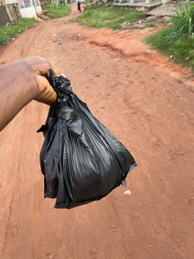 The image shows a person's hand holding a black plastic bag filled with something, which is tied off at the top. The hand appears to be of a person with dark skin. The background is a dirt road or path with reddish-brown soil, which suggests a rural or semi-urban environment. Some green grass and vegetation are visible along the side of the path, and in the distance, there are some buildings that look like houses, suggesting a residential area. The overall scene appears to be outdoors. There is no visible text in the image. The lighting suggests it could be daytime, but the exact time of day is not clear. The context provided indicates the location is Edopa, Nigeria.