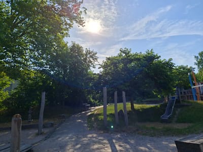 The image depicts a playground area on a sunny day, likely located in Sinzheim, Germany. The scene is dominated by lush green trees casting dappled shadows on a paved pathway made of cobblestones. In the foreground, several rough-hewn wooden posts stand vertically. To the right, a metal slide descends from a wooden play structure, part of which is visible with blue and orange accents. A few large rocks are scattered on the grassy area around the playground equipment. In the middle ground, a path curves through the trees, leading towards a simple wooden bench. The sky above is a vibrant blue with wispy white clouds and the sun shining brightly, creating lens flare effects. The overall impression is one of a serene outdoor recreational space.