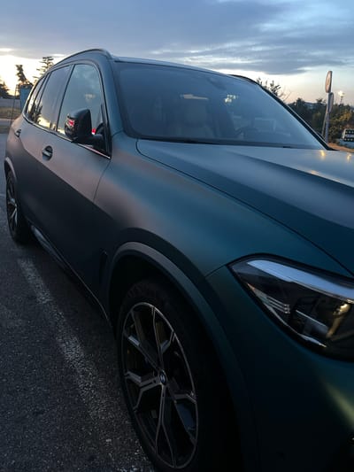 The image shows a matte teal BMW X5 parked on an asphalt road with white markings, likely a parking lot or roadside. The sky is overcast with hints of sunset colors visible on the horizon. The car's exterior is the primary focus, with its sleek lines and modern design emphasized by the low-angle shot. The background features trees and some distant buildings, including a sign that appears to be in Slovenian, suggesting the location is Šempeter pri Gorici, Slovenia. The lighting indicates it is either dawn or dusk. There are no people visible in the image.