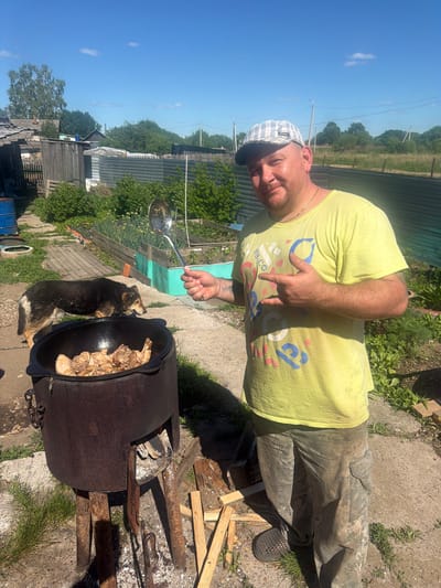 In Valdgeym, Russia, a man wearing a plaid cap, a light yellow t-shirt, and camouflage pants stands in a yard. He holds a large metal spoon in his right hand, pointing with his left index finger towards a large black cauldron filled with meat, which is being cooked over an open fire. To the left of the cauldron, a dog with black and brown fur looks towards the cooking meat. The background shows a rural setting with wooden fences, green vegetation, and a blue sky with scattered clouds.