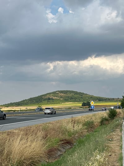 The media file displays a roadside scene with a highway running through a rural landscape. The perspective is from the side of the road, looking down the highway which curves to the right. On the left side of the highway, there is a steep, green, tree-covered hillside. The right side of the highway is bordered by dry, tall grass and some sparse vegetation. The sky is heavily clouded, with patches of brighter sky indicating sunlight filtering through.

Several vehicles are visible on the highway, moving in the same direction. Closer to the foreground, a silver SUV and a dark-colored car are traveling. Further down the road, other vehicles are seen, including a white van and a dark car. There are road signs present, including a yellow diamond-shaped sign with a black border and a blue circular sign with a white arrow pointing left, indicating a turn or lane change. Another triangular sign is visible further down the road.

The time of day appears to be daytime, given the lighting conditions and the visibility of the landscape. The weather seems to be overcast or partly cloudy, with the possibility of impending rain due to the dark clouds. The location is identified as Drazhevo, Bulgaria, suggesting a rural or semi-rural setting in Eastern Europe. The scene conveys a sense of travel and movement along a road. No people are distinctly visible, but their presence is implied by the vehicles. The overall mood is neutral, reflecting a typical travel scene.
