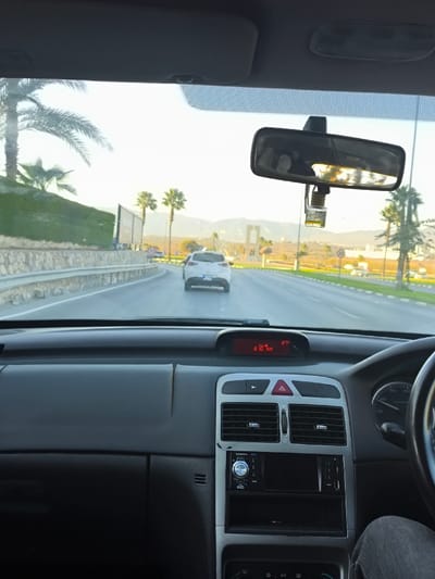 The image provides a forensic view from the passenger seat of a right-hand drive vehicle, looking forward on a multi-lane asphalt road in Gonyeli, Northern Cyprus. The vehicle's dark gray dashboard is prominent, featuring a central digital display indicating "210 km" and "27°C," an aftermarket car stereo head unit, and a red triangular hazard light button. A rearview mirror hangs centrally, reflecting bright sunlight, with a yellow-bottled air freshener attached beneath it. The driver's leg, clad in blue jeans, is partially visible on the far right.

Ahead on the road, a white Mazda hatchback, noticeably covered in dust, maintains distance from the camera vehicle. To the left, a low stone wall supports a metal guardrail, beyond which a green-foliage-covered embankment with several palm trees and a billboard structure is visible. In the mid-distance, centrally positioned, the distinct Gonyeli Martyrs Monument stands on what appears to be a roundabout or central island. Additional palm trees are scattered along the roadside, and a range of mountains stretches across the far background under a clear, bright sky, indicative of either morning or late afternoon light.