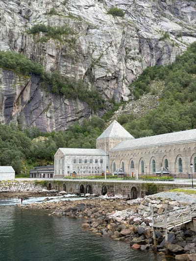 In Glomfjord, Norway, a large, historic power station complex is situated at the base of a steep, rugged cliff face. The water in the foreground appears to be a river or fjord, with rocky shores and turbulent rapids. A lone figure, dressed in red, stands on the rocks in the water, facing the rapids.
The power station itself is a substantial, stone structure with a long, arched facade and a prominent, pyramid-shaped roof element. Adjacent to it is a smaller, lighter-colored building. The cliff behind the structures is covered in dense green trees, with exposed grey rock faces showing evidence of erosion and stratification. A metal bridge and walkway extend over the rocky bank towards the water. The sky is overcast, contributing to the muted tones of the scene.