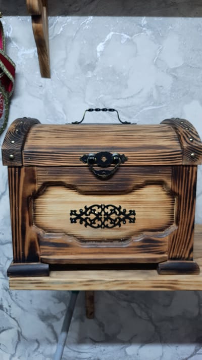A wooden chest with a decorative metal handle and ornate filigree is displayed on a wooden shelf. The chest has a rustic, burnt wood finish and is adorned with metal studs along its lid. The background features a wall with a marbled pattern. A decorative ribbon in red and gold is partially visible on the left side of the image. The scene is indoors, likely in a residential setting. There are no people present in the image.