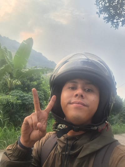 A young man is wearing a black motorcycle helmet and a brown jacket. He is making a peace sign with his right hand. The man is positioned in the foreground. Behind him is a lush green landscape consisting of a mix of trees and bushes, with a hill in the background. The sky is overcast. The location is in Kecamatan Margaasih, Indonesia, at the coordinates -6.926 latitude and 107.536 longitude.