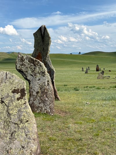 The image depicts a field of standing stones under a cloudy blue sky. The foreground features three large, weathered stones covered in lichen. In the midground, numerous smaller standing stones are scattered across a green, grassy plain. The background consists of rolling green hills, with a single tree visible on one of the hills. The scene appears to be an archaeological or historical site, likely in a rural, open landscape. The location is identified as Сафронов, Russia.