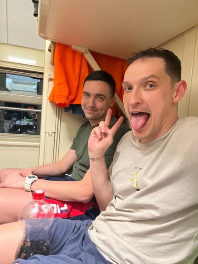 Two men are inside a train carriage. One man is making a face with his tongue out, while the other man is making a peace sign. They are sitting close together. The train carriage contains bedding, a window, and a small table. There is a plastic bottle of soda on the lap of one of the men. There is no evidence to support the claim that the location is in the city of Kizner, Russia.