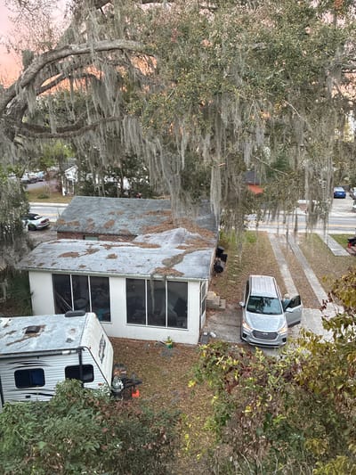 An elevated, overhead view depicts a residential property and street scene in Gainesville, United States of America, under soft, diffused lighting with a faint orange hue on the distant horizon, indicative of sunrise or sunset.

The upper portion and foreground are dominated by a large, mature tree heavily draped with Spanish moss (Tillandsia usneoides) and exhibiting green foliage. Below the tree, a single-story house is visible. The main section of the dwelling features a dark, shingled roof substantially covered in fallen leaves. An attached white sunroom or screened porch extension, equipped with large windows/screens, has a flatter, lighter-colored roof also bearing scattered leaves. To the left of the sunroom, a white travel trailer or recreational vehicle (RV) with a weathered roof and a propane tank on its hitch is parked on the ground.

A silver minivan or large SUV is parked on a paved driveway to the right of the sunroom. Its front passenger door is open. A single individual, characterized by light-colored hair and wearing a light-colored top, is partially visible near the open passenger door, in the process of exiting or standing adjacent to the vehicle. The ground surrounding the structures consists of sparse dry grass, bare soil, and fallen leaves.

In the background, a paved street runs horizontally. A white convertible sports car with green-accented wheels is parked on the street to the left, positioned behind the main house. Further to the right on the street, a blue sedan is parked. Additional residential structures and trees are discernible lining the street in the distance.