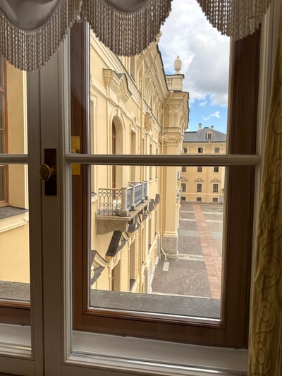 This is a photograph taken from inside a building, looking out through a large window. The window has wooden frames and is partially obscured by a decorative valance with fringe at the top and a golden curtain on the right side.
Through the window, we see an architectural scene. The main subject is a large, pale yellow building with ornate detailing, including classical columns, arched windows, and a decorative balcony with a metal railing. The architecture suggests a historical or imperial style, consistent with locations in Strelna, Russia, particularly the Konstantinovsky Palace.
Beyond the immediate building, another, darker-roofed building is visible in the distance, also appearing to be part of a grand complex. The ground visible is a courtyard or plaza paved with patterned bricks. The sky above is partly cloudy with patches of blue. The time of day appears to be daytime, likely late morning or early afternoon, given the lighting conditions. No people are visible in the frame. The overall impression is one of looking out onto a historic courtyard from a well-appointed interior.