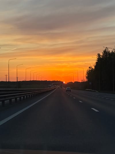 The image shows a highway at sunset. The location is likely Khmelevka, Russia, as indicated by the presence of Russian signage. The highway has multiple lanes and is separated by a guardrail. Cars are visible in the distance. Streetlights line the side of the road, casting shadows across the landscape. The sky is a gradient of colors, ranging from light gray to vibrant orange. Trees can be seen along the road.