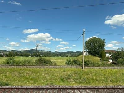 The image presents a static outdoor scene captured in Wetzikon (ZH), Switzerland, on a bright, sunny day, likely during spring or summer given the lush green vegetation. The sky, occupying the upper third of the frame, is a clear, vibrant blue with scattered, fluffy white cumulus clouds.
In the immediate foreground, two parallel sets of train tracks are visible. The tracks consist of dark brown rails resting on grey ballast, which is interspersed with green grass and weeds. The tracks recede slightly from the viewer's perspective.
Beyond the foreground tracks, a wide expanse of verdant green field stretches across the midground. This field is densely covered with grass and features scattered bushes and taller herbaceous plants. Several dark utility poles, part of an overhead electrical catenary system for trains, punctuate the landscape, supporting multiple dark wires that traverse the sky horizontally. In the distance within the midground, a paved road is visible, with several light-colored vehicles (likely cars) appearing as small, indistinct shapes. Rolling hills, densely covered with trees and some lighter areas of fields, form the horizon in the background.
To the far right of the image, residential structures are present. A substantial white building with a reddish-brown tiled roof is partially visible, nestled behind trees and a grassy mound. On top of this mound, a smaller, simple white structure with a grey roof stands prominently. No people, active subjects, or dynamic actions are depicted within the frame.