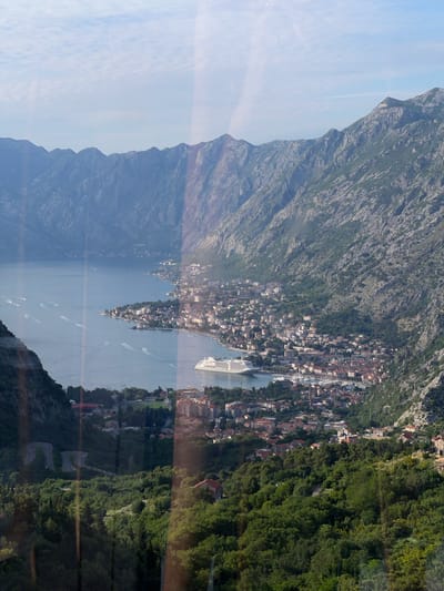 This detailed image provides a high-angle, panoramic view of a coastal landscape, identified as Čavori, Montenegro. The scene encompasses a large, calm bay flanked by imposing, rugged mountains under a hazy light blue sky with faint cloud patterns.
Prominent in the background and midground are steep, rocky mountains, predominantly grey-blue with scattered green vegetation on their lower slopes. The vast blue-grey water of the bay, likely the Bay of Kotor, occupies the central and left portions of the frame. Numerous small boats are visible on the water, leaving distinct white wakes, indicating movement.
Along the right side of the bay, a dense coastal town extends, characterized by numerous buildings with distinctive red-tiled roofs. This settlement stretches from the water's edge up the lower mountain slopes. A large, white multi-deck cruise ship is conspicuously docked or anchored near the town's waterfront in the mid-right. A harbor area with smaller vessels is also discernable within the town.
In the foreground and lower left, dense green foliage and trees cover the slopes. A winding road with visible switchbacks is cut into the verdant hillside in the bottom left quarter of the image. Vertical light streaks and subtle reflections are present across the image, suggesting it was photographed through glass or a similar transparent medium. There are no individuals explicitly visible or identifiable.