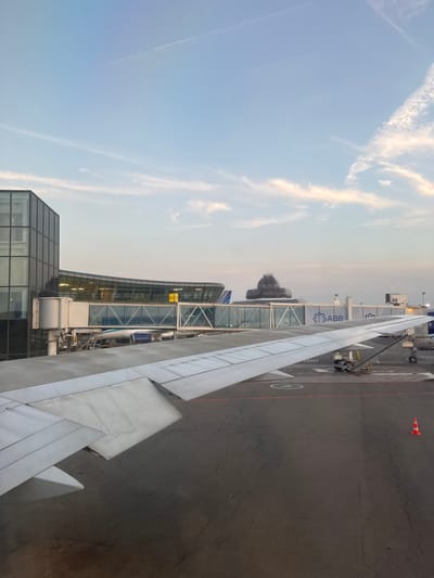 This image captures a scene at an airport, likely during the late afternoon or early evening given the soft, warm lighting on the clouds. The perspective is from inside an airplane, looking out the window. The prominent subject is the wing of the aircraft, its metallic surface stretching across the lower right portion of the frame.

Beyond the wing, an airport terminal building with modern architecture is visible. It features large glass windows that reflect the sky. A jet bridge connects the terminal to an unseen aircraft. Several signs are visible on the jet bridge, including one with the prominent blue logo and text "ABB". Another sign displays "A02 A2". An orange and white traffic cone sits on the tarmac, a common sight at airports. The tarmac itself appears weathered, with visible cracks. In the distance, the rounded structure of a control tower or a dome-like building is discernible.

The sky is a pale blue, with wispy clouds tinged with the warm hues of the setting sun. The overall impression is one of quiet activity at an airport, possibly during boarding or disembarking. There are no people clearly visible in the frame. The location is identified as Khazar Raion, Azerbaijan.