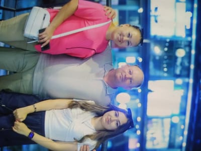 The image shows three individuals, a woman and a man standing, with a young girl sitting. The woman in the foreground is wearing a pink shirt and beige pants and holding a black smartphone. She has a white purse on her left shoulder. The man is in the middle, wearing a light gray shirt. The young girl in the foreground is wearing a white shirt and dark pants. She has a watch on her left wrist and bracelets on her right wrist. The background appears to be an indoor setting with bright blue and white lights. The image is oriented vertically, with the heads of the individuals at the top and their feet at the bottom. This scene does not appear to be specific to the city of Birlik, Uzbekistan, and there are no identifying landmarks or contextual clues to suggest this location.