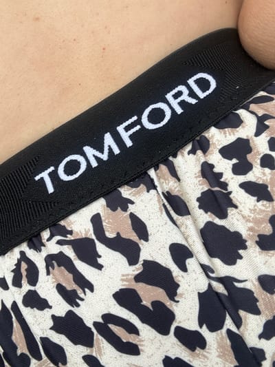 A close-up, eye-level shot shows the elastic waistband of a pair of leopard-print underwear. The black waistband has the white text "TOM FORD" printed on it in capital letters. The underwear fabric is gathered and folded, creating a textured surface. The background consists of a person's skin, appearing pale and lightly speckled with small reddish marks. The image does not provide any information about the city of Ayyrtau, Kazakhstan, or any contextual details that would place it within that location.