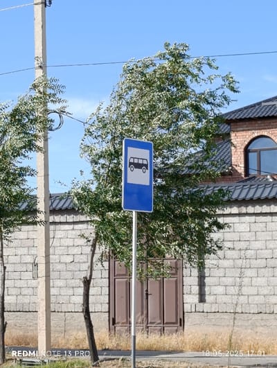 A blue and white bus stop sign with a silhouette of a bus is mounted to a white pole. The sign is visible against a brick wall and a large tree with green leaves. The pole is supported by a cement base. The brick wall is adjacent to a brown wooden door. There is dry grass growing at the base of the wall. The photo was taken in Türkistan qalası, Kazakhstan, at coordinates 43.304, 68.319. The image was captured on May 18, 2025, at 5:01 PM.