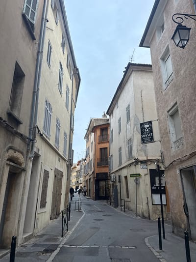 An alleyway is lined with aged buildings of varying heights and textures, their windows often covered by shutters. The sky is overcast. A dark sign, "Sunset Musique Billard", hangs above. Smaller signs and lighting fixtures add to the street's eclectic character. The narrow paved street slopes downwards, with figures walking towards the deeper end, creating a sense of perspective.