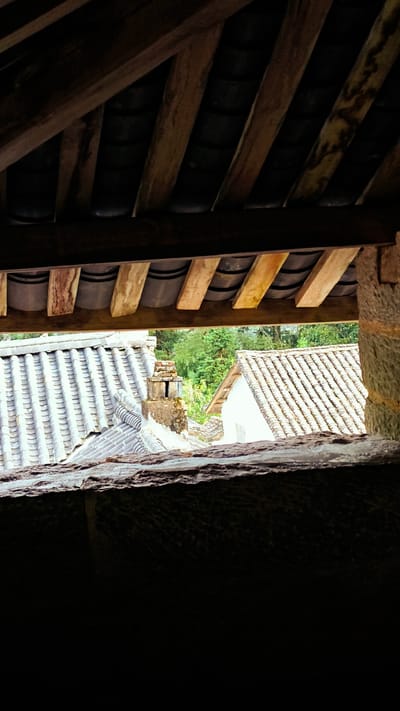 The image captures a partial view of tiled rooftops in Sà Phìn Commune, Vietnam, as seen through a dark opening. The foreground is dominated by dark shadowed stone walls. Above, the rough-hewn wooden beams of a ceiling with dark, round tiles between the beams create a layered effect. Beyond the opening, two visible rooftops with traditional gray tile roofing are prominent. One has a brick chimney. The background shows lush green vegetation.