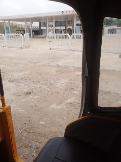 The image shows a view from inside a yellow auto-rickshaw looking out towards a building with a white sign that reads "MERITL" and a white metal fence. The auto-rickshaw is parked in a gravel parking lot, and the building is in the background. There is a single piece of white paper lying on the ground, and the sky is overcast.  The scene is located in the city of Abuja, Federal Capital Territory, Nigeria, at latitude 8.955 and longitude 7.552. There are no people present.