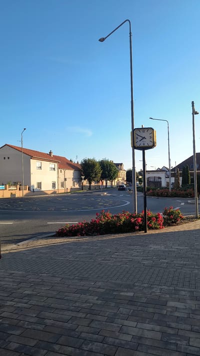The image depicts a street scene in Rudná, Czechia, with a clear blue sky. In the foreground, dark gray paving stones form the sidewalk. A cluster of vibrant red roses is planted along the edge of the sidewalk, bordering a street. The street itself features yellow speed bumps arranged in a circular pattern, indicating a roundabout or traffic calming measure.

In the middle ground, a square clock with a yellow frame is mounted on a tall, dark pole. The clock face shows the time as approximately 9:10. Behind the clock, a row of houses with red tiled roofs lines the street. To the left, a light-colored building with multiple windows and a reddish-brown tiled roof is visible. Further down the street, trees with lush green foliage stand, and more buildings with similar architecture can be seen.

A dark-colored car is driving on the street. A lamppost with a bent arm is positioned to the left of the clock. Another lamppost, taller and thinner, stands to the right of the clock. The environment appears to be well-maintained, with elements suggesting a residential or mixed-use area within the town. The lighting suggests it is daytime, possibly late afternoon, given the long shadows cast by the lampposts.