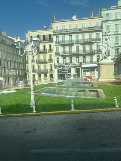 The image depicts a bright, sunny day in Toulon, France, likely in a public square. In the foreground, a section of asphalt road with a yellow curb is visible. Beyond the curb lies a well-maintained green lawn dotted with white bollards. A tiered fountain with cascading water is the central feature of the lawn.

Dominating the background are several multi-story buildings with classical architecture. These buildings feature light-colored facades, numerous windows with shutters, and decorative balconies with iron railings. A prominent white street lamp with multiple lights stands in the middle ground, adding to the urban ambiance.

To the right of the fountain, a large stone statue stands on a pedestal. The statue appears to be of two figures, one pointing outwards. In front of the buildings, several businesses are identifiable, including "Guston" a hair salon, and "POSTAL" a postal service. A red car is parked in front of one of the businesses. The sky above is a clear, vibrant blue.