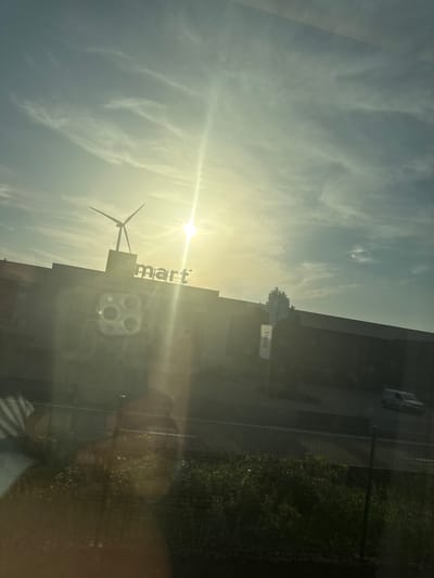 The image shows a building with the word "mart" visible on its facade, illuminated by the sun. A wind turbine is positioned to the left of the building. The sun is bright and appears to be either rising or setting, casting a lens flare across the image. The sky is partly cloudy with wispy clouds. In the background, a street is visible with a white car driving on it. To the right of the building, there is a patch of green vegetation. Reflections are visible on the surface from which the photo was taken, obscuring parts of the scene. The overall impression is that of an outdoor scene, possibly a commercial area, with a focus on the "mart" building and the surrounding environment.