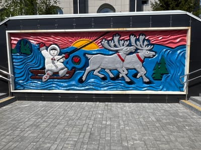 The image shows a bas-relief mural on the exterior of an underpass in Neryungri, Russia. The mural depicts an indigenous person in a white parka and fur-lined hood, seated in a wooden sled being pulled by two reindeer across a body of water. The scene is framed by a red sky with a yellow sun and is set against a backdrop of blue waves, green trees on the left, and a single green tree on the right. The person in the sled is holding reins and appears to be guiding the reindeer. The reindeer are grey with large antlers and red collars. The artwork is colorful and appears to be made of painted concrete or plaster. In the foreground, there is a paved walkway made of grey rectangular stones. Stairs and a metal handrail are visible on the right side of the image. The overall scene conveys a theme of Arctic life and travel.