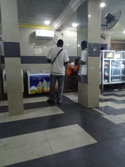 The image shows a view inside a commercial space, likely a shop or restaurant, in Ovwian, Nigeria, at coordinates 5.503 latitude, 5.799 longitude. There are two men standing, one facing the camera and the other facing away, both with their backs to a large white freezer that has a sticker advertising ice cream. The freezer sits beside a large pillar, painted a light beige color, and a third man is seated in front of the freezer, partially obscured by the pillar. The man facing the camera is wearing a white t-shirt, dark pants, and a dark crossbody bag. The man with his back to the camera is wearing a white t-shirt and dark pants. The seated man is wearing an orange shirt and dark pants. The floor is a black and white checkerboard tile pattern. There is a ceiling-mounted air conditioner in the top left corner of the frame and a ceiling fan in the upper right corner. The space is well-lit with overhead fluorescent lights. Behind the men, there is a clear glass display case filled with various items, likely food, and behind the case, there is a metal shelving unit. The image is taken at eye level.