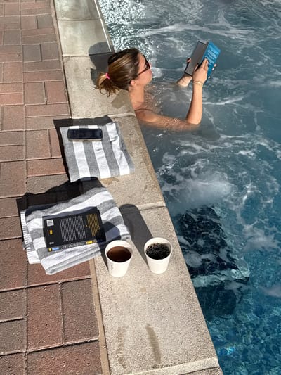 An overhead shot captures a woman in a jacuzzi, reading. Two striped towels rest poolside: one holds a book and the other, a phone. Two white cups, one seemingly filled with coffee, sit nearby on the concrete ledge separating the water from the brick-paved area. The jacuzzi water is frothy and reflects light. The scene suggests a leisurely, relaxed setting.