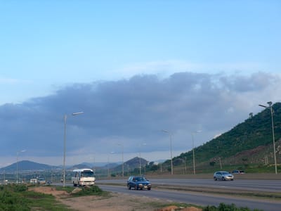 This is a sideways, landscape-oriented photograph taken from a road. The image shows a road with several vehicles on it, including a white bus, a blue car, and another white vehicle. The road appears to be an highway or a major road. On the left side of the road, there is a grassy hill with trees. On the right side of the road, there are more grassy areas and distant mountains. The sky is cloudy with a mix of gray and blue hues. Several lampposts are visible along the side of the road. The image appears to be taken during the daytime, likely in the late afternoon or early evening given the lighting and the presence of clouds. The overall scene suggests a travel or transportation context in a somewhat rural or semi-urban environment. There are no discernible people in the image, and no visible text. The photograph is oriented with the top of the image on the left, giving a sense of the scene being viewed from the side or if the camera was tilted.