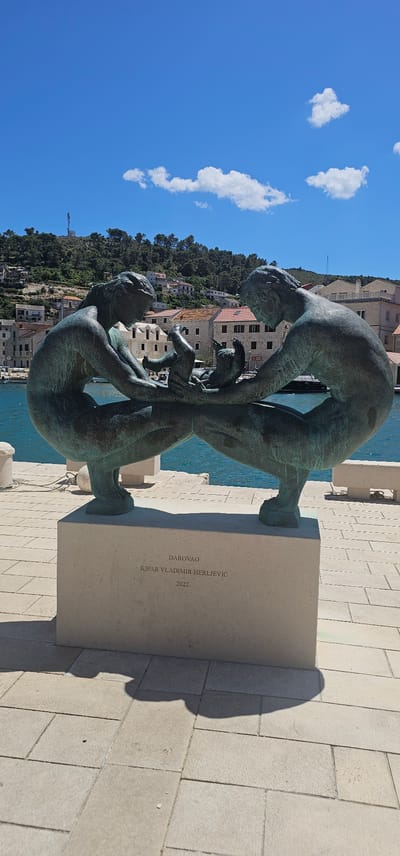 In Općina Pučišća, Croatia, a bronze sculpture depicting two adults and two infants is positioned on a stone pedestal by the water. The sculpture is dark green in color and shows the figures in a crouched, embracing posture, with the infants between them. The stone pedestal has an inscription that reads "DAROVAO Kipar VLADIMIR HERLJEVIĆ 2022." In the background, there is a body of blue water, a stone promenade, and a hillside covered with trees and buildings with red tiled roofs, under a bright blue sky with scattered white clouds.