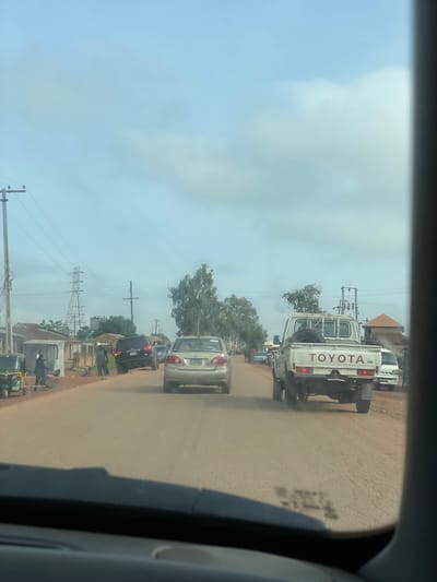 The image captures a scene on a dusty road in Kassa Market, Nigeria. The perspective is from inside a vehicle, looking out. A white Toyota pickup truck is the most prominent vehicle, seen from the rear, driving away from the viewer. A spare tire is mounted on the back of the truck. To the left of the Toyota, a silver sedan is also traveling in the same direction. Further ahead, a black SUV is visible, parked or moving slowly. On the left side of the road, several small structures that appear to be market stalls or homes are present. A green auto-rickshaw is parked near one of these structures. Several individuals are visible on the roadside, some standing and others seemingly engaged in activity. Electrical poles and overhead wires are prominent features of the environment, indicating the presence of infrastructure. Trees and vegetation line the sides of the road, with a larger cluster of trees in the mid-ground. The sky is overcast with clouds. The overall impression is that of a busy, somewhat dusty, and well-trafficked market area in Nigeria.