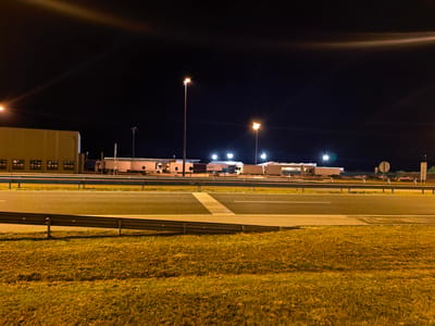 The image depicts an industrial area at night, likely near a highway, in Obrežje, Slovenia. The scene is illuminated by artificial lights from street lamps and building lights, casting a warm yellow glow on the environment.

The primary subjects are several large buildings and a row of parked trucks, suggesting a logistics or distribution hub. The buildings are long and rectangular with multiple windows. The trucks are mostly white, with some appearing to be semi-trailers. A few cars are also visible, parked in front of the buildings or on the roadside. A highway runs in the foreground, separated by a metal guardrail from a grassy verge. The sky is dark, indicating nighttime, and there are some bright light flares and streaks from the artificial lighting, characteristic of a long-exposure shot or direct light sources. There are no people visible in the frame.

The activity taking place appears to be passive, with vehicles and buildings in a state of rest. The overall mood is quiet and functional, typical of an industrial site after operational hours. The time of day is clearly night. The weather is not explicitly discernible, but the dry appearance of the grass suggests it might not be raining.

A notable detail is text on one of the trucks, which appears to read "EUROPOOR". There are also road signs visible in the distance, including a circular white sign with a black border. The location context is provided as Obrežje, Slovenia.